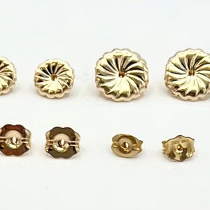 May include: A set of six gold-toned earring backs. The set includes two larger, round backs with a swirl design, two smaller, round backs with a smooth surface, and two smaller, square backs with a smooth surface.