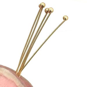 May include: Close-up of several gold-colored head pins. Each pin features a small, round ball at the top and a long, slender shaft. The pins are arranged together, with some overlapping, against a white background. These pins are ideal for jewelry making and crafting.