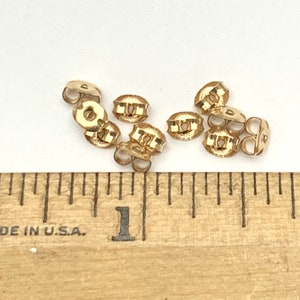May include: A collection of small gold-colored metal earring backs positioned above a wooden ruler that shows measurements in inches. The earring backs are circular with a central opening and two curved wings for securing an earring post.
