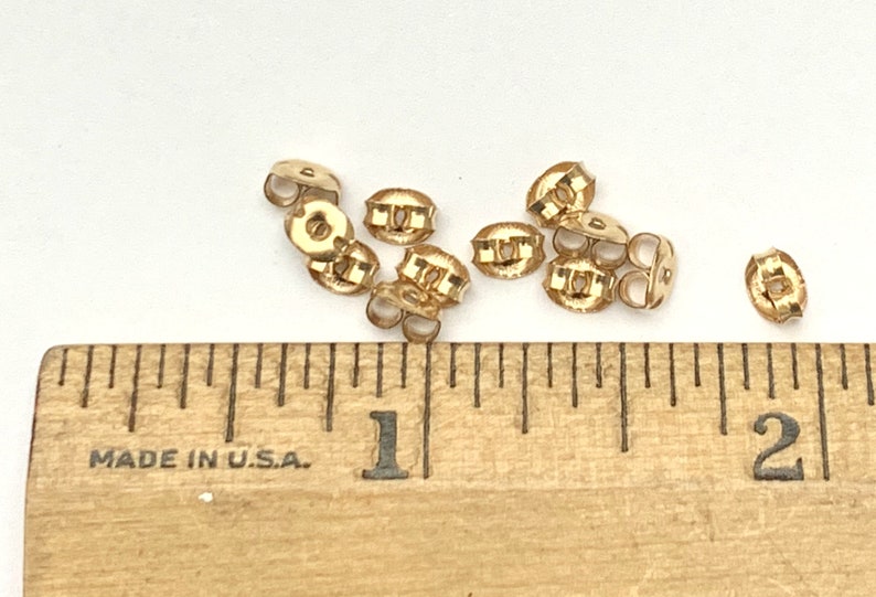 May include: A collection of small gold-colored metal earring backs positioned above a wooden ruler that shows measurements in inches. The earring backs are circular with a central opening and two curved wings for securing an earring post.
