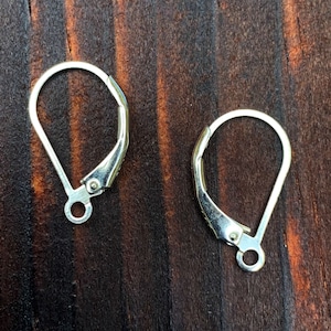 May include: Two silver earring hooks, also known as French hooks, are shown on a dark brown wooden surface. The hooks are made of metal and have a smooth, polished finish.
