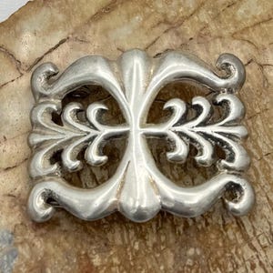 May include: A silver-toned, ornate, cross-shaped pendant with intricate floral designs. The pendant is made of metal and has a polished finish.