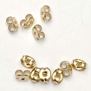 May include: A collection of ten gold-toned metal earring backs, each featuring a small circular center hole and two curved wings designed to grip an earring post securely. These small jewelry components are arranged in two loose groups.