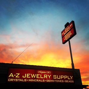 May include: A rectangular illuminated sign for Majesta M's A-Z Jewelry Supply, Crystals, Minerals, Gemstones, Beads, mounted on a building and a tall pole against a vibrant orange, yellow, and blue sunset sky, displaying the business name and items.