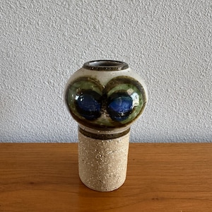 May include: A ceramic vase with a cylindrical base featuring a speckled beige texture. The upper portion is bulbous and smooth, decorated with two prominent circular motifs in shades of deep blue, forest green, and dark brown glaze.