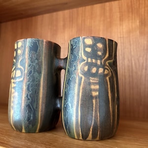 May include: A double-chambered ceramic vessel featuring a dark, mottled glaze in shades of charcoal, deep green, and bronze. The exterior surface displays vertical, textured patterns with lighter, yellowish-brown etched geometric motifs.