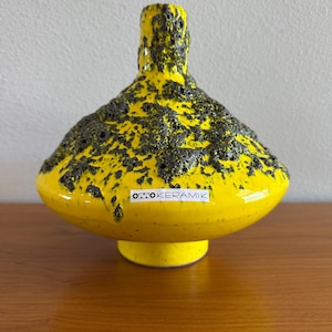 May include: A bright yellow ceramic vase with a wide, rounded body and a narrow neck. The upper portion features a thick, dark grey, textured lava-like glaze overlay. A white label on the front displays the black text OTTO KERAMIK.