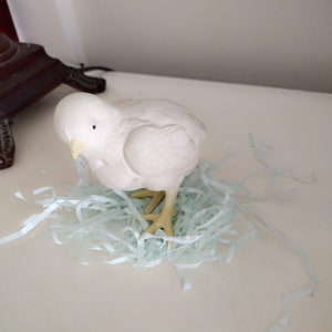 May include: A white ceramic chick figurine sitting in a nest of light blue shredded paper.
