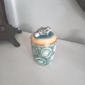 Pode incluir: Uma caixa de bugigangas decorativa de cerâmica com uma figura de sapo na tampa. A caixa é cilíndrica com um padrão verde e branco, uma borda dourada e um topo laranja claro. O sapo é verde com manchas marrons e detalhes vermelhos.