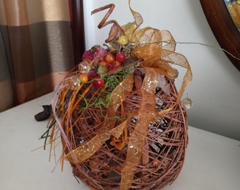 May include: A decorative, spherical autumn-themed ornament. The base is made of woven brown twigs, adorned with orange and gold ribbons, berries, and decorative elements. The ornament is designed to resemble a pumpkin.