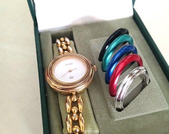 May include: A gold-toned Gucci watch with a white face and a gold band, presented in a green box. Includes a set of interchangeable colored bezels in black, teal, blue, red, and silver.