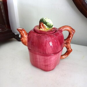 Peut inclure: Théière en céramique en forme de pomme rouge, avec une feuille verte et jaune sur le dessus. La poignée et le bec sont bruns, ressemblant à une branche. La théière est posée sur une surface blanche.