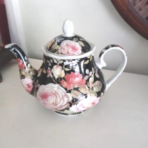 以下が含まれることがあります： ピンクと白のバラのフローラルパターンが施された、蓋付きの黒いセラミックティーポット。 ティーポットは、湾曲した注ぎ口とハンドル、白いバラのつぼみのつまみがあります。 お茶好きのための装飾品。