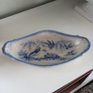 May include: Oval-shaped, white ceramic dish with a blue bird and floral design. The rim is trimmed with a blue scalloped border. The dish has a slightly raised edge and is likely vintage or antique.