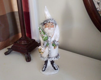 May include: White Santa Claus figurine with a green Christmas tree in his hand. He is wearing a white fur coat with silver tinsel trim and a white hat.