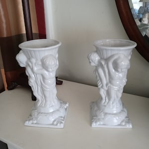 Könnte beinhalten: Zwei weiße Keramikvasen mit Cherubfiguren, die die Vase halten. Die Vasen haben einen quadratischen Sockel mit einem floralen Design.