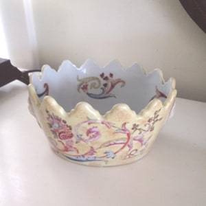 Puede incluir: Un cuenco decorativo con borde festoneado, exterior crema e interior blanco. Presenta diseños florales y foliares en rosa, rojo, azul y verde. El cuenco es probablemente de porcelana o cerámica.