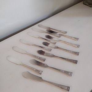 May include: A collection of silver butter knives arranged on a white surface. The knives have a rounded blade and a rectangular handle design. The blades are shiny, reflecting light, and the handles have a decorative pattern.