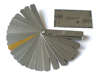 Könnte beinhalten: Ein Saiten-Action-Lineal aus Metall mit einem Fächer aus Metallmesslehren zur Messung der Saitenhöhe an Musikinstrumenten. Das Lineal ist in 64stel Zoll und Millimetern gekennzeichnet.