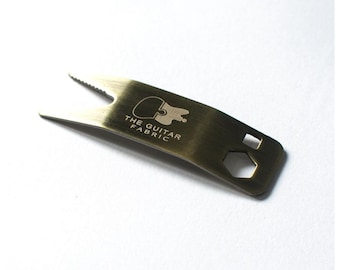 Könnte beinhalten: Ein goldfarbenes Metall-Gitarrenwerkzeug mit einem "THE GUITAR FABRIC"-Logo und mehreren Aussparungen für verschiedene Funktionen.