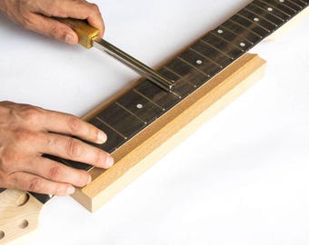 Könnte beinhalten: Nahaufnahme eines Gitarrenhalses, der mit einem Metallwerkzeug bearbeitet wird. Der Hals besteht aus hellem Holz mit dunklen Bünden und weißen Punkteinlagen. Das Werkzeug hat einen Holzgriff und eine Metallklinge. Der Hintergrund ist weiß.