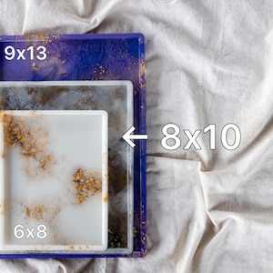 May include: Three resin trays in different sizes, stacked on top of each other. The top tray is purple with gold glitter, the middle tray is white with gold flecks, and the bottom tray is white with gold flecks. The trays are labeled with their dimensions: 9x13, 8x10, and 6x8.