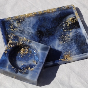May include: A set of two blue and white resin trays with gold flecks. The larger tray is rectangular and the smaller tray is square with a round indentation in the centre.