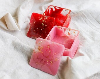 May include: Three square resin boxes in shades of red and pink, with gold flakes embedded. One box is open, revealing its interior. The boxes are set on a white, textured fabric, creating a soft, diffused background.