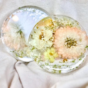 Puede incluir: Dos posavasos circulares de resina con flores secas incrustadas en tonos rosa pálido, crema y amarillo suave. La resina transparente y pulida conserva los delicados pétalos y el follaje verde, creando una superficie lisa y transparente.