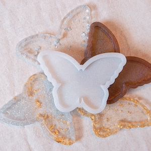 May include: Three butterfly-shaped resin trays, one white, one brown, and one clear with gold glitter. The white tray is a silicone mold.