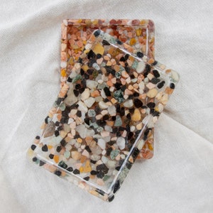 Peut inclure: Deux plateaux rectangulaires en résine remplis de petits cailloux colorés. Les plateaux sont transparents avec un bord, et les pierres sont dans les tons de noir, blanc, jaune, orange et vert. Un plateau recouvre partiellement l'autre.