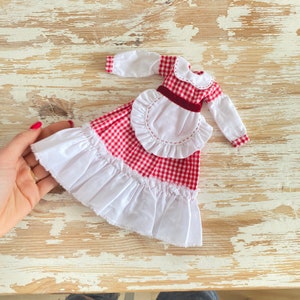 May include: A doll dress featuring a red and white gingham pattern with long white sleeves, a white collar, and a white ruffled apron. The dress is accented with a dark red velvet ribbon at the waist and a wide white ruffled hem at the bottom.