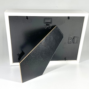 May include: White picture frame with a black mat and a black easel stand on the back. The frame has a white border and a black backing.