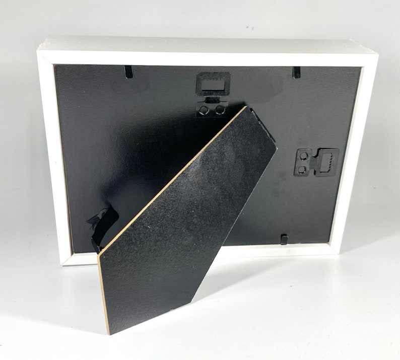May include: White picture frame with a black mat and a black easel stand on the back. The frame has a white border and a black backing.