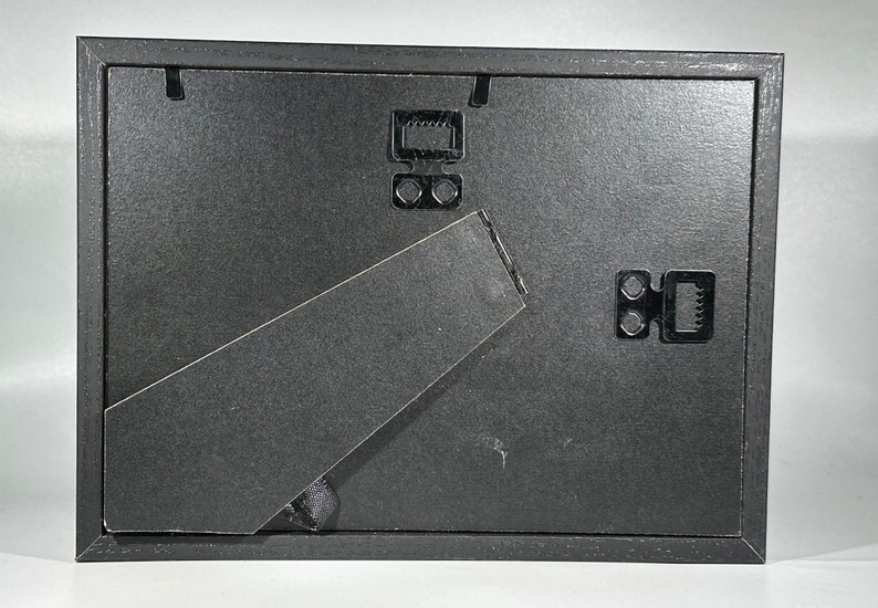 May include: Black picture frame with a black easel stand on the back. The frame has two black metal hanging hooks on the back.