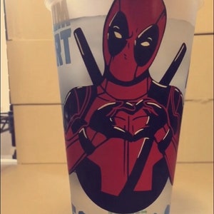 May include: A white plastic cup with a red and black Deadpool character design. The character is making a heart shape with his hands. The text "Deadpool" is printed in blue on the cup.