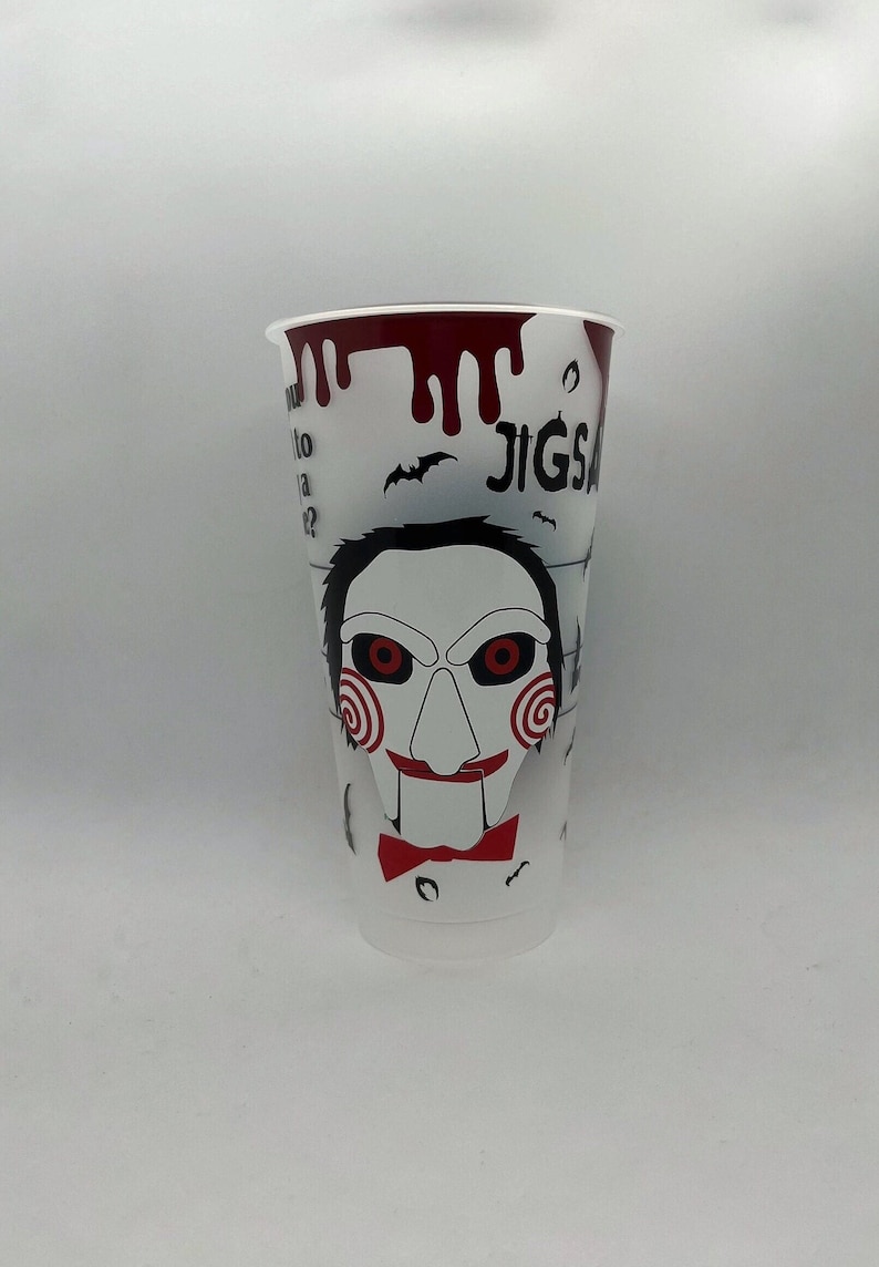 May include: White plastic cup with a red, black, and white graphic of Jigsaw from the Saw movie franchise. The graphic includes the text "Jigsaw" and a red and black spiral design.
