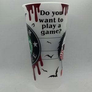 May include: White plastic cup with a black and red design. The cup has a Starbucks logo and the text "Do you want to play a game?" with a dripping blood effect. The cup also features a black and white illustration of a character from the horror movie "Saw".