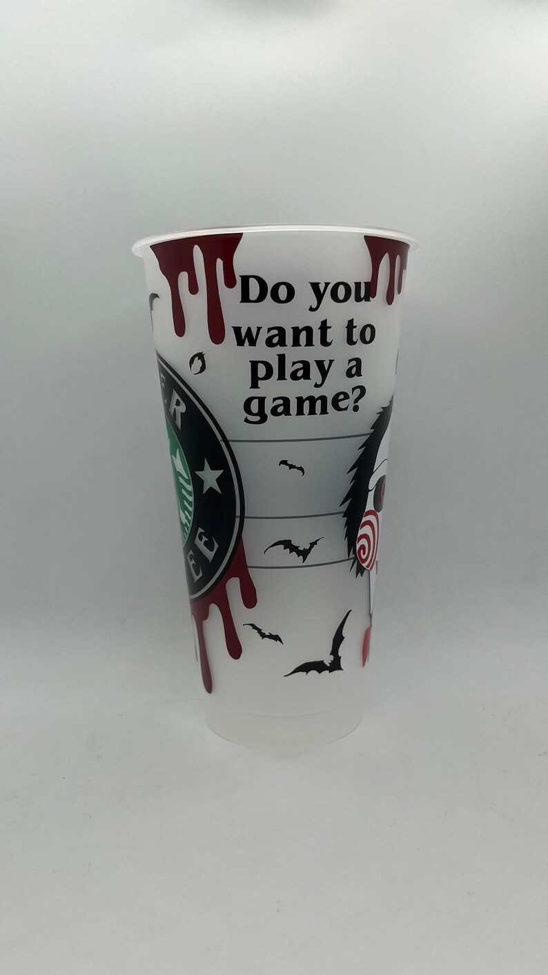 May include: White plastic cup with a black and red design. The cup has a Starbucks logo and the text "Do you want to play a game?" with a dripping blood effect. The cup also features a black and white illustration of a character from the horror movie "Saw".