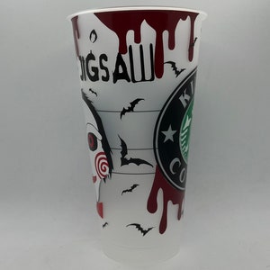 May include: A white plastic cup with a black and red design. The cup features a graphic of a character from the horror movie "Saw" with the text "Jigsaw" and a Starbucks logo. The cup is decorated with red blood splatters and black bats.