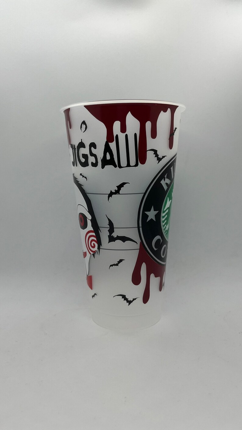 May include: A white plastic cup with a black and red design. The cup features a graphic of a character from the horror movie "Saw" with the text "Jigsaw" and a Starbucks logo. The cup is decorated with red blood splatters and black bats.
