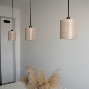 May include: Three wooden pendant lights hang from the ceiling above a white table with a vase of dried pampas grass. The lights are made of light wood and have a cylindrical shape.