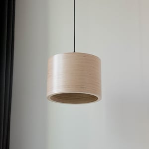 May include: A light wood pendant light fixture with a cylindrical shade hangs from a black cord against a white wall.
