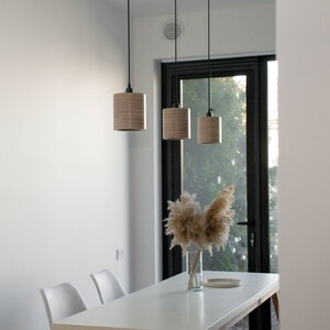 May include: Three wooden pendant lights hang over a white table with two white chairs. A vase with dried pampas grass sits on the table in front of a large window.