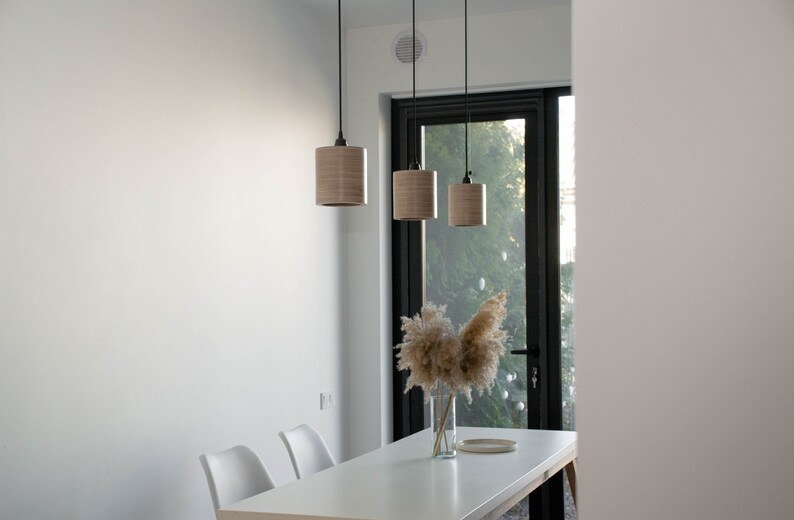 May include: Three wooden pendant lights hang over a white table with two white chairs. A vase with dried pampas grass sits on the table in front of a large window.