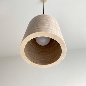 May include: A wooden pendant light fixture with a natural wood finish. The light fixture has a cylindrical shape with a circular opening at the bottom. The light fixture is hanging from a black cord.