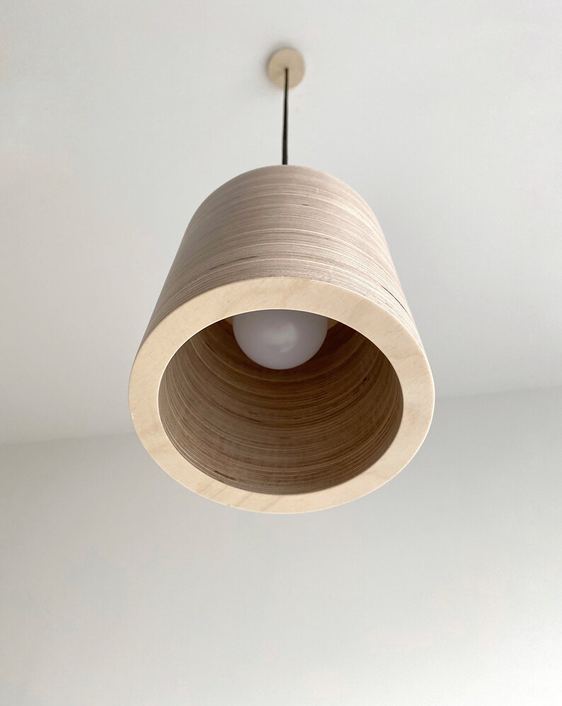 May include: A wooden pendant light fixture with a natural wood finish. The light fixture has a cylindrical shape with a circular opening at the bottom. The light fixture is hanging from a black cord.
