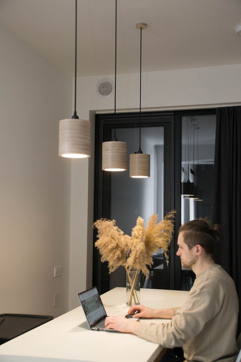 May include: A person sits at a white table with a laptop, working. Three wooden pendant lights hang above the table. A vase with dried pampas grass sits on the table.