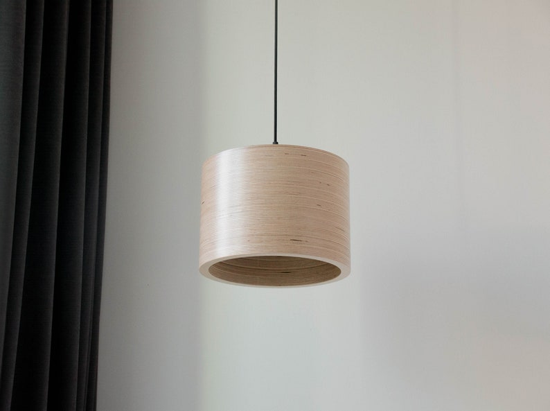 May include: A light wood pendant light fixture with a cylindrical shade hangs from a black cord against a white wall.