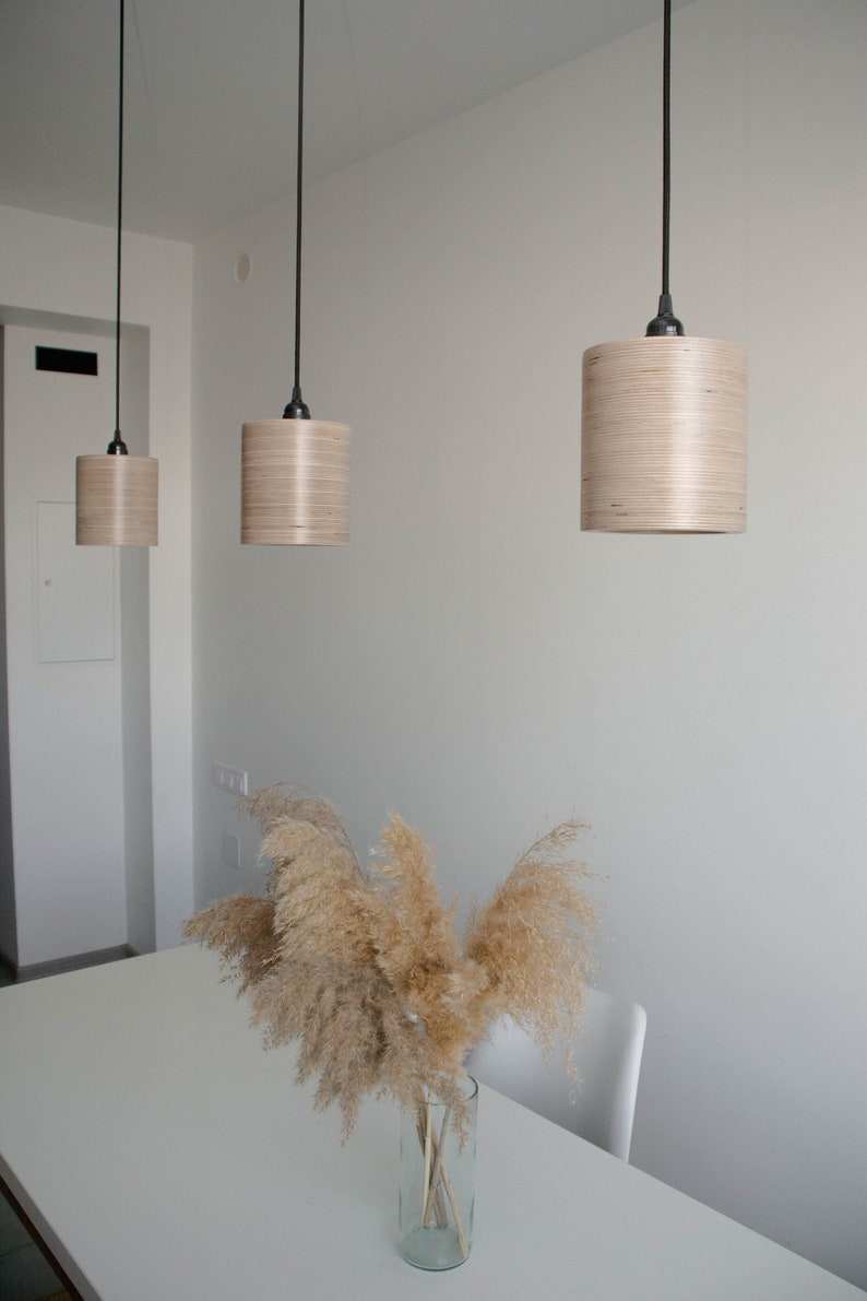 May include: Three wooden pendant lights hang from the ceiling above a white table with a vase of dried pampas grass. The lights are made of light wood and have a cylindrical shape.